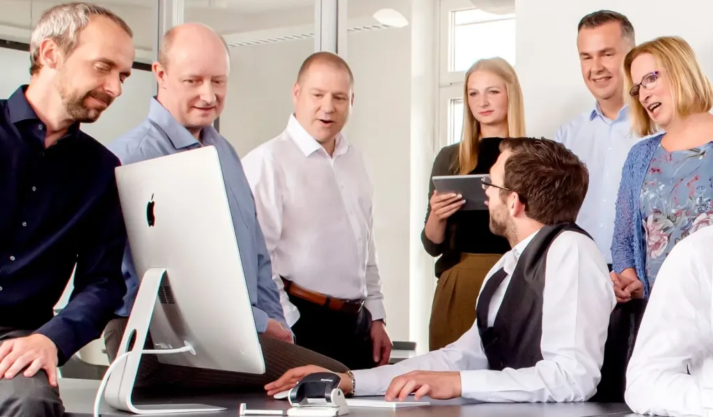 Das Team vom IT Dienstleister Berlin bizIT in einem modernen Büro. Im Vordergrund sitzt ein Mann mit Weste am Schreibtisch vor einem großen Computerbildschirm, während die anderen Personen stehend miteinander sprechen und lachen.